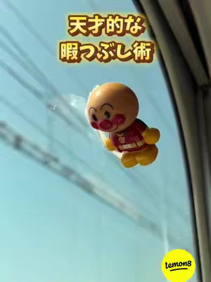 An Anpanman toy with a suction cup is stuck to a window, with a blue sky visible outside. Text overlays the image in vibrant yellow.