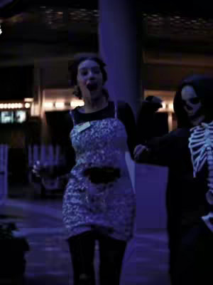 A costumed female with an open mouth is running, to the right of the frame, and a person in a black skeleton costume follows her. Blue light surrounds the frame and the background is out of focus.