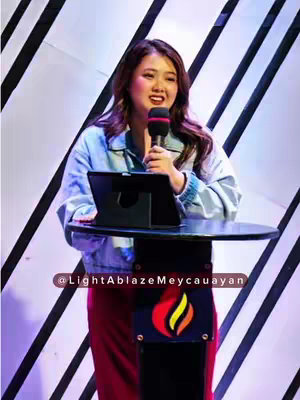 A woman with long brown hair and wearing a light blue jacket stands at a podium, speaking into a microphone. A tablet rests on the podium. The background has diagonal white stripes. The bottom of the podium has a logo with flames.