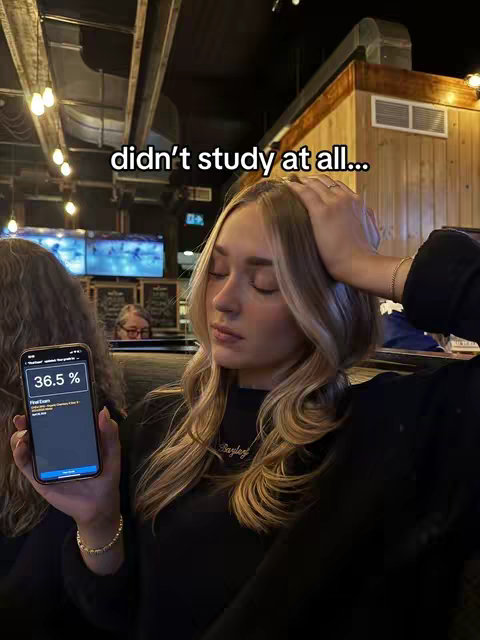 A woman with long blonde hair is holding her head with one hand and a smartphone with the other. The phone screen displays a percentage. The background suggests an indoor setting.