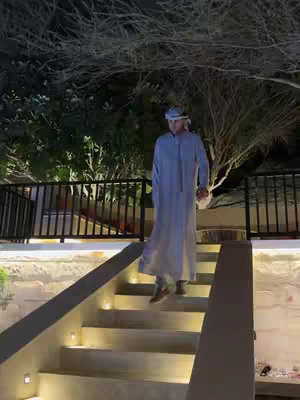 A man in traditional Arabic clothing walks down a lit staircase at night, with trees and a dark sky in the background, suggesting a Middle Eastern or Arabian Gulf location.