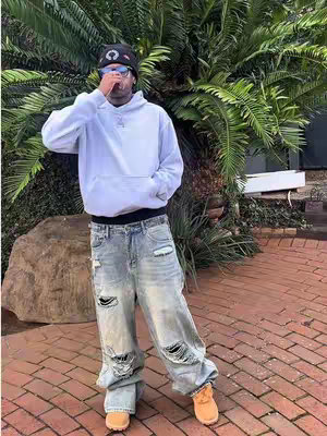 A man in a white hoodie, ripped jeans, and a black beanie poses outdoors. He wears sunglasses, and a brick pathway and lush greenery form the background.