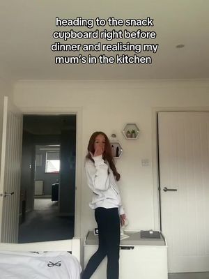 A young woman with long red hair stands in a room, blowing a kiss towards the camera. She's wearing a white hoodie and black pants. The background includes a white door, a bed, and a room with a door open with a view of a hallway.