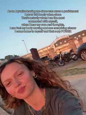 A selfie-style shot featuring a young woman with long, wavy hair, looking at the camera with a slight smile. The background includes motorcycles, a car, and a building. The image has a warm, natural lighting. Text overlayed on the image provides context.