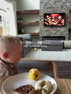 A young child sits at a table with a plate of food in front of them, looking to the side. The background includes a living room with a TV and fireplace.