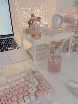 A well-lit, aesthetically pleasing desk setup with a laptop on the left, a keyboard in the foreground, and decorative items on a white desk shelf. The keyboard has pink and white keycaps. The overall color scheme is pastel and soft, with a focus on productivity and aesthetics. A pink drink with a cute sticker is on the tabletop.
