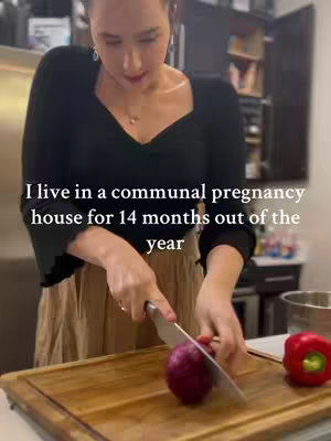 A woman in a black top and tan skirt is in a kitchen, holding a knife and cutting a red onion on a wooden cutting board. A red pepper sits nearby. Text overlay indicates she lives in a communal pregnancy house.