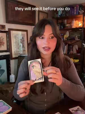 A woman with long brown hair is holding up a tarot card while looking directly at the camera. She is in a room with framed pictures on the wall and a bookshelf in the background.