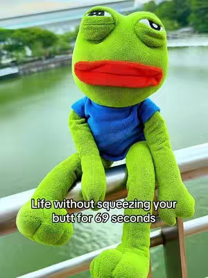 A plush frog with green fur and red lips is sitting on a metal railing. Behind the frog, there is a green water body and trees in the background and blue sky can be seen in the background.