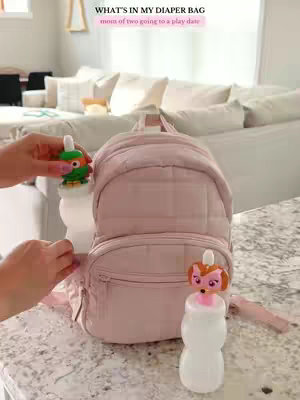 The cover image shows a light pink mini backpack prominently displayed on a white countertop, flanked by two baby bottles with whimsical animal character toppers. A hand is placing a bottle near the backpack. The background is a blurred living room scene with a white couch, adding context suggestive of a home environment. Text overlays state 'WHAT'S IN MY DIAPER BAG' and 'mom of two going to a play date', indicating the video's content focus.