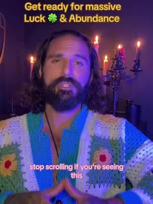 A medium shot of a man with a beard and long hair, wearing a textured, colorful sweater. Text overlays at the top say, "Get ready for massive Luck & Abundance", while text at the bottom says, "stop scrolling if you're seeing this". In the background are candles and a decorative candle holder. The lighting has a purple hue.