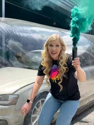 A woman with long, blonde wavy hair smiles widely while leaning against a silver car. She holds a green smoke flare, which is emitting a large cloud of green smoke. The background is a sunny outdoor scene.