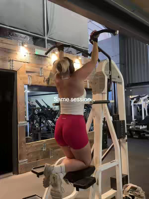 A female is shown from the knees up, actively doing pull-ups on a workout machine. She is wearing pink shorts and a white top. It's a medium shot in a well-lit gym setting, with workout equipment visible in the background.
