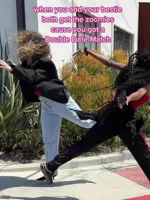 Two young women are captured mid-action in an outdoor setting. The foreground includes two figures, one in a black top and pants in a running pose, and the other in a black jacket and blue jeans, leaning away in a similar energetic stance. Behind them, greenery and a building with windows create a backdrop under a clear sky.