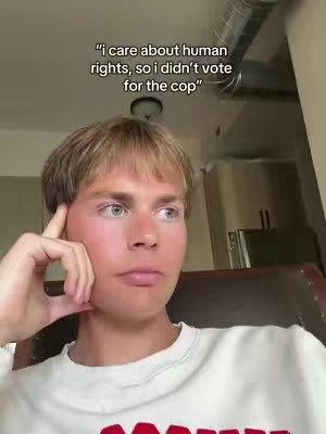 The cover image features a young man with light skin, short blond hair, and blue eyes. He is wearing a white sweatshirt and appears to be in a room with natural light and visible architectural details like a metal vent and a wooden chair. Text overlay at the top quotes: "i care about human rights, so i didn't vote for the cop". He's looking off to the side, with his index finger touching his cheek as if he's contemplating something. The image is framed from the chest up, highlighting the man's face and expression.