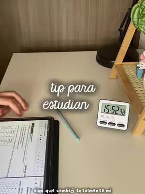 A clean, well-lit, top-down shot of a desk setup. A tablet with a partially visible screen displaying what appears to be a study guide is front and center. A light blue stylus rests next to the tablet. To the right sits a white timer and a partially visible plant. The background appears to be a plain beige wall.