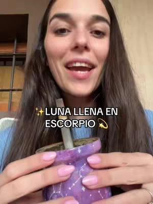 A woman with long, dark hair is shown in a selfie-style video. The top half of the image is dominated by her face, with her mouth open in a speaking position. She's holding a purple vessel with a straw inside, and her pink fingernails are visible. The background is slightly blurred, with sunlight streaming through a window.