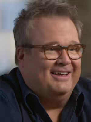 A medium shot of Eric Stonestreet, wearing glasses and a dark blue shirt, smiling. The background is out of focus, suggesting an interview setting.