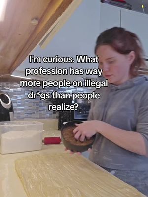 A person stands in a kitchen, the lower half of the image showing a counter with ingredients and an unfinished pastry, and a woman using two hands to hold a tray that appears to be inside of a pan.