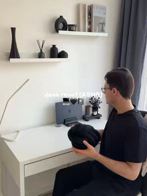 A person is seen from the chest up, standing near a white desk. On the desk is a laptop, notebook, and various small decorative items. There are shelves with minimalist decor displayed on the white wall behind the desk. The person is holding a black object.