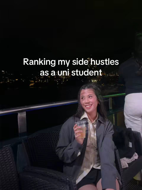 A young woman with medium-length dark hair, wearing a dark grey blazer over a cream-colored shirt and black shorts, smiles with a clear beverage in a clear plastic cup. She is seated on a dark wicker chair in front of a cityscape at night. Text overlays read "Ranking my side hustles as a uni student".