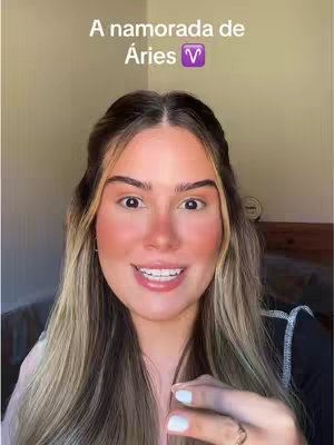 The cover image features a young woman with long blonde hair, smiling directly at the camera. She has fair skin, with blush applied to her cheeks, and is wearing a black top. The background is a neutral, light-colored wall. Text overlayed at the top reads, "A namorada de Áries," with the Aries zodiac symbol. A ring with a gold band is visible on her right hand. The composition is a close-up, conveying a friendly and engaging tone.