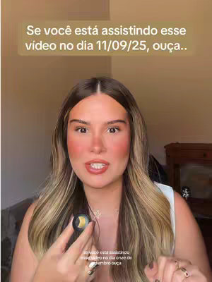 A woman with long light brown hair and light makeup is in a selfie video. She is holding a small circular object in her hand. The background is somewhat blurred beyond the camera.