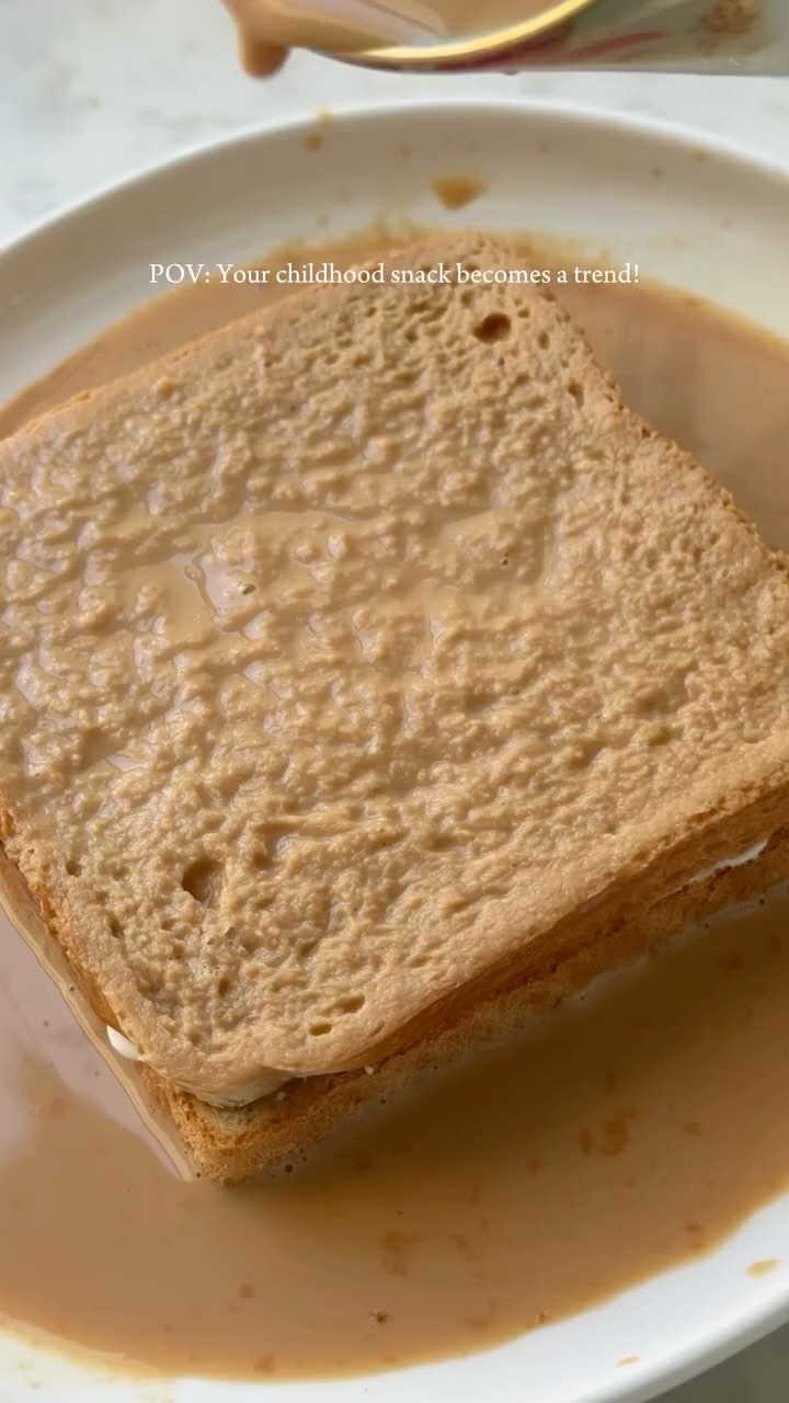 The image shows a close-up of a piece of bread submerged in a light brown liquid, likely a sauce or milk-based mixture. The bread is a light brown color. At the top of the image, a hand can be seen pouring the liquid over the bread from a container. The composition is simple, with the bread as the central focus, and the liquid enveloping the bread, suggesting a food-related theme. 