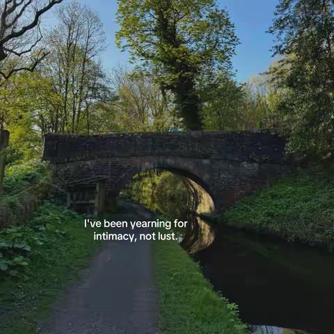 A scenic shot of an old stone bridge arching over a canal, surrounded by lush greenery and trees. The water reflects the bridge and the sky, and text overlayed at the bottom of the image that says 'I've been yearning for intimacy, not lust.'