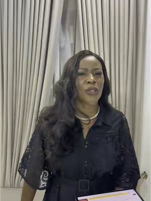 A medium shot features a woman speaking, framed from the chest up. She is positioned slightly off-center, facing the camera with a serene expression. She is wearing a black dress that has a decorative pattern, and a pearl necklace. Her long, dark hair is styled with loose waves that cascade over her shoulders. The background consists of vertical, light-colored curtains that suggest an indoor setting.