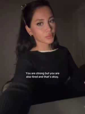 A woman with dark hair in a black sweater is looking towards the camera. Text overlayed reads: 'You are strong but you are also tired and that's okay.' The image has low lighting, and the background is blurred.
