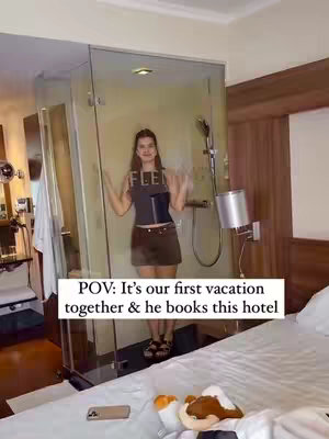 The cover image shows a woman standing in a hotel room, posing in front of a glass shower. In the foreground, a bed with pillows and some items is partially visible. The woman is wearing a black tank top and a brown skirt. Text overlayed on the image reads: 'POV: It's our first vacation together & he books this hotel'.