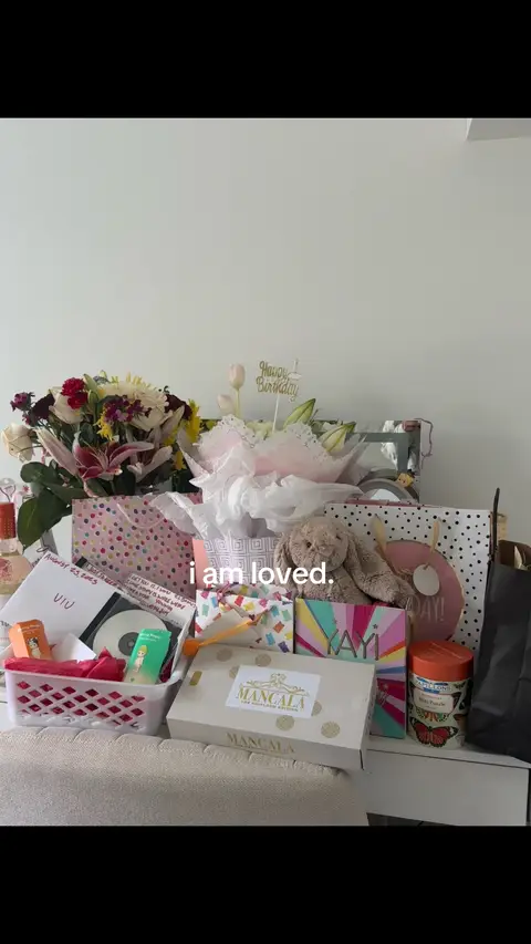 The image shows a collection of birthday gifts arranged on a surface. There are various wrapped presents, flowers, a stuffed animal, and a cake topper that reads "Happy Birthday". The text overlayed says "i am loved."