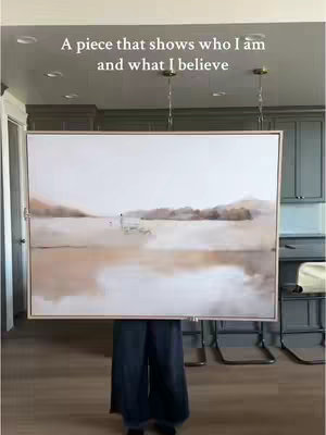A woman stands in front of an art piece. The artwork is a landscape painting in neutral tones. The background includes a kitchen scene.