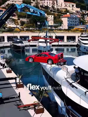 A red Ferrari F40 is being lifted by a crane from a marina onto a superyacht. The scene is set at a harbor with luxury yachts and buildings in the background with bright sunlight and blue water.