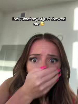 A selfie featuring a young woman with wide eyes and a hand covering her mouth in a surprised expression. The woman's long, brown hair frames her face, and she wears bright pink nail polish. Text above reads, "look what my bf just showed me". The background is blurred, suggesting an interior setting.