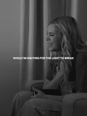A black and white image shows a woman seated and looking to her right with a gentle smile. She appears to be in a chair with her hands resting on her lap. In the background is what looks like curtains, with the lighting being dim. Text overlayed on the image reads "WHILE I'M WAITING FOR THE LIGHT TO BREAK".