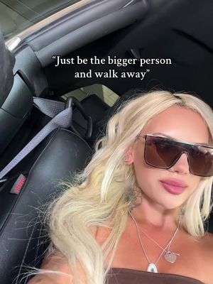 A selfie-style image taken from inside a car. A woman with long, wavy, blonde hair and large sunglasses is in the foreground, with the car interior visible around her and the driver's seat and headrest in the background. The upper portion of the image displays text overlay.