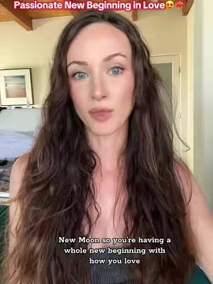 A woman with long, wavy brown hair is centered in the frame, looking directly at the camera. She has fair skin, blue eyes, and is wearing a dark top. Above her head, at the top of the frame, there is text that says, 'Passionate New Beginning in Love'. Below her, at the bottom, there is text again, starting from the left, saying, 'New Moon so you're having a' and ending with 'how you love'. The background shows the interior of what appears to be a house, with a light brown wall on either side and a wooden beam across the ceiling.