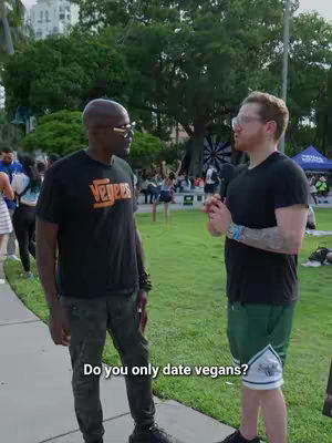 Two men are in an outdoor setting, engaged in a conversation. The man to the left, dressed in a black t-shirt with white text and camouflage-style pants, wears dark sunglasses and has a serious expression. The man on the right, wearing a black t-shirt and green shorts, holds a microphone and appears to be the interviewer. A crowd of people is visible in the background, and the setting looks like a park or outdoor event space.