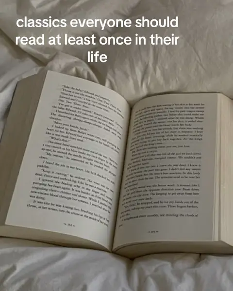 An open book rests on a bed, its pages filled with text, suggesting a scene of reading in a comfortable setting. The book appears well-worn, with a soft-focus background of a white blanket.