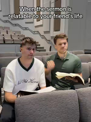 Two young men are seated in what appears to be a church or auditorium, with rows of gray seats visible. The man on the left is wearing a white sweater with a black v-neck and text on it, holding a paper. The man on the right, holding a book, has a slight smile as though conveying inside information.