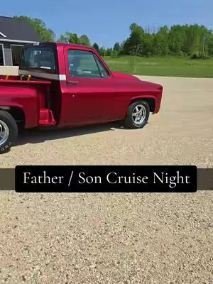 A red pickup truck is parked on gravel, against a backdrop of green trees and a blue sky. The truck has a black text bar over the lower portion of the image. The overall lighting is bright with a natural look.