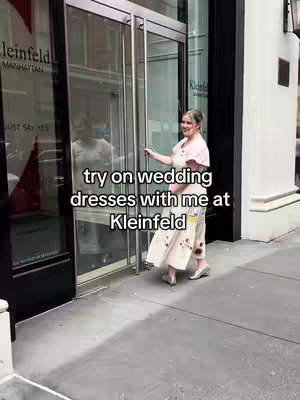 A woman in a wedding dress is entering a building with glass doors. The text overlay says 'try on wedding dresses with me at Kleinfeld'. The scene is brightly lit, and the woman is smiling.