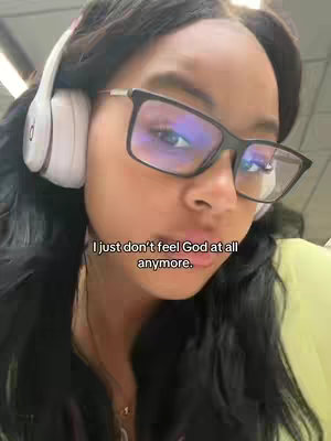 A selfie-style shot of a person wearing black-framed glasses and white headphones. The person has dark hair and is against a blurry background. A text overlay stating "I just don't feel God at all anymore."