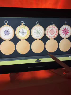 An angled view of a black screen showing a game with six stylized compasses with varying internal designs, each above a yellow semi-circle. A hand holding a pencil is visible in the lower-right corner of the frame.