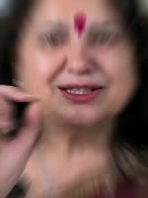 A close-up shot of a woman's face with an out-of-focus effect. She is smiling, and a bindi adorns her forehead. The blurred background suggests a soft focus, highlighting her expression.