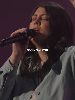 A medium shot features a woman with dark hair singing into a microphone. She is wearing a pale blue top, and the background is blurred with a purple hue. The overlay text 'YOU'RE ALL I WANT' is positioned near the bottom.