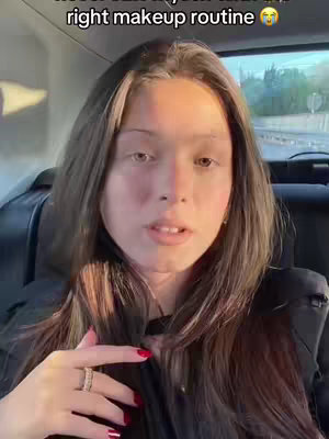 A woman with long brown hair is seated in the back of a car, looking at the camera. She has fair skin, is wearing a black top, and has red nail polish. The background is blurred, suggesting movement or a busy environment.