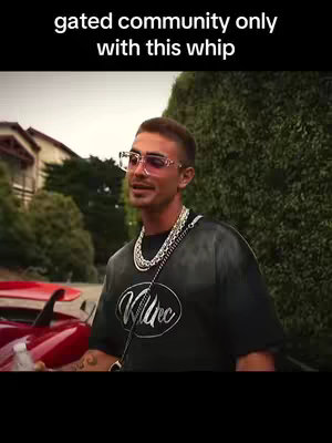 A man wearing glasses and a chain necklace is standing outside a building. The man is wearing a black t-shirt with white text where appears the word "Killtec".
The scene in a gated community.